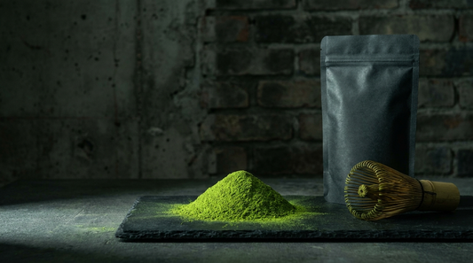 Vivid green ceremonial matcha powder with a bamboo whisk and matte black pouch on dark stone against an industrial brick background